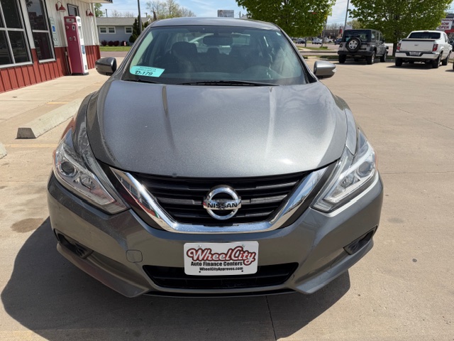 Front view of a gray Nissan sedan at a dealership, Wheel City Auto Finance Centers license frame, parking lot and buildings in the background.