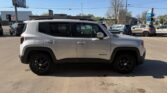 Side view of a silver Jeep Renegade with black wheels parked in a dealership lot, with other cars and buildings in the background.