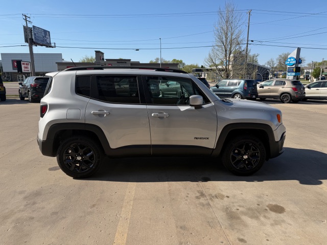 Side view of a silver Jeep Renegade with black wheels parked in a dealership lot, with other cars and buildings in the background.