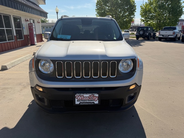 Front view of a white Jeep SUV in a car dealership lot, showing the seven-slot grille and round headlights; license plate reads Wheel City nearby.