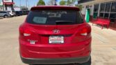 Rear view of a red Hyundai Tucson parked in a car dealership lot; D-179 sticker on the rear window and a Wheel City Auto Financing Center plate, with a building and red bench in the background.
