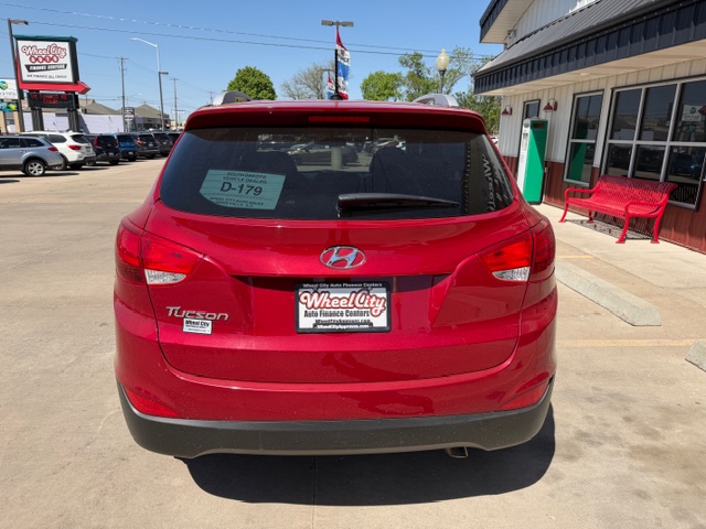 Rear view of a red Hyundai Tucson parked in a car dealership lot; D-179 sticker on the rear window and a Wheel City Auto Financing Center plate, with a building and red bench in the background.