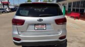 Rear view of a silver Kia SUV parked in a dealership lot, featuring a Wheel City license plate frame and a red bench beside a storefront.
