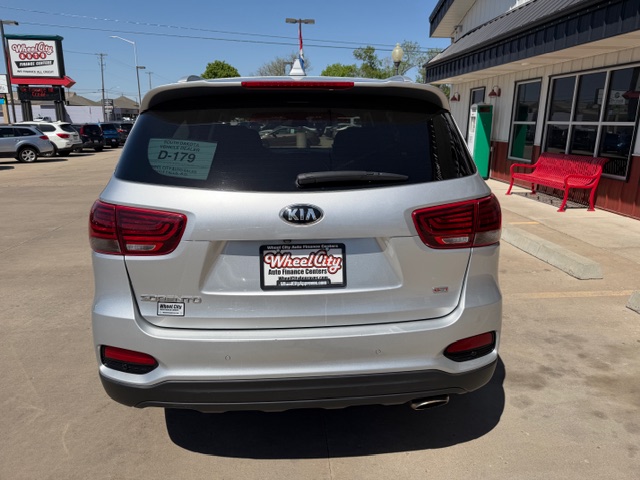 Rear view of a silver Kia SUV parked in a dealership lot, featuring a Wheel City license plate frame and a red bench beside a storefront.