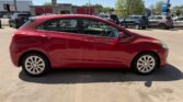 Red hatchback car parked in a lot, side view with dealership buildings in the background.