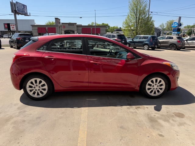 Red hatchback car parked in a lot, side view with dealership buildings in the background.