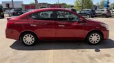 Side view of a red four-door sedan parked in a sunny dealership lot with other cars in the background, visible parking lines.