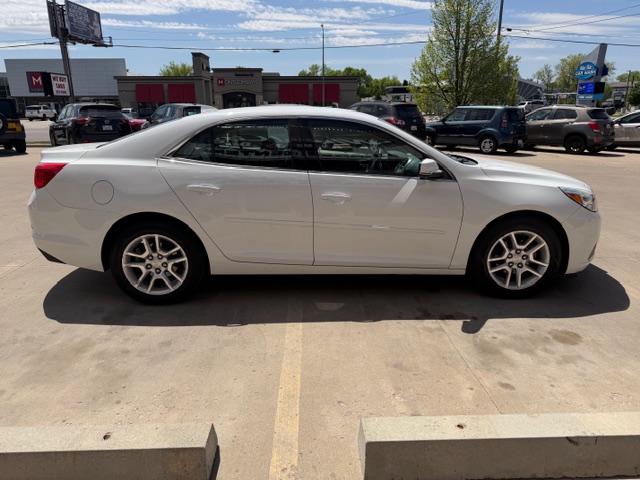 White four-door sedan photographed from the side in a parking lot, with other vehicles and storefronts in the background.