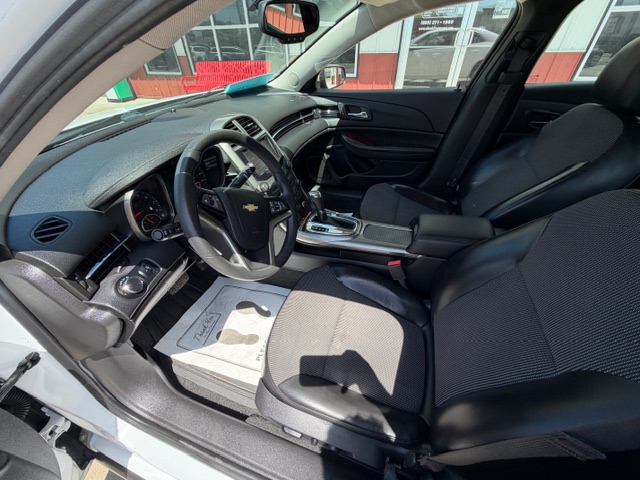 Interior of a white Chevrolet sedan: driver’s seat, front passenger seat, steering wheel with Chevy logo, dashboard, and center console; driver's door is open.