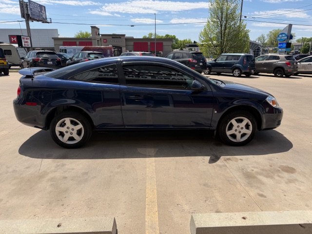 Side view of a dark blue two-door coupe parked in a parking lot, with other cars and a row of buildings in the background.