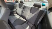 Backseat of a four-door car with gray patterned upholstery and three seat belts visible, sunlit from the side window.