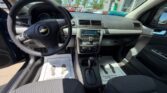 Driver's perspective of a Chevrolet car interior: steering wheel, center console, gear shifter, and front seats with floor mats visible through open driver door.