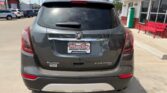 Rear view of a gray Buick Encore SUV parked in a car dealership lot, with Wheel City Auto Finance Centers license plate frame and red benches outside a building.