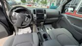 Interior of a gray pickup truck from the back seat, showing the steering wheel, center console with radio and controls, gear shifter, and front seats; dealership lot visible through the windshield.