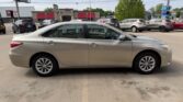 Beige four-door sedan shown in a side profile parked in a car dealership lot with other vehicles and storefronts in the background on a sunny day.