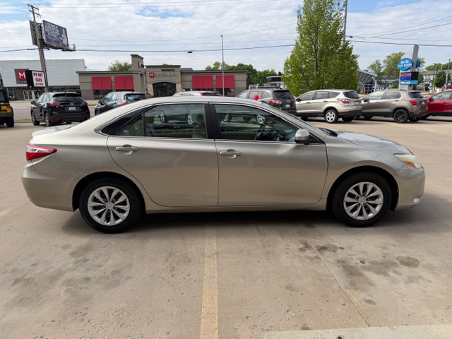 Beige four-door sedan shown in a side profile parked in a car dealership lot with other vehicles and storefronts in the background on a sunny day.