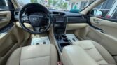 Inside a beige Toyota car: steering wheel, center touchscreen, gear shifter, and beige seats; dealership lot visible through the windows.