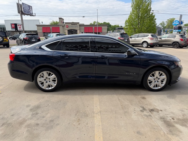 Side view of a dark blue Chevrolet Impala sedan parked in a parking lot, with tinted windows and alloy wheels, storefronts in the background.