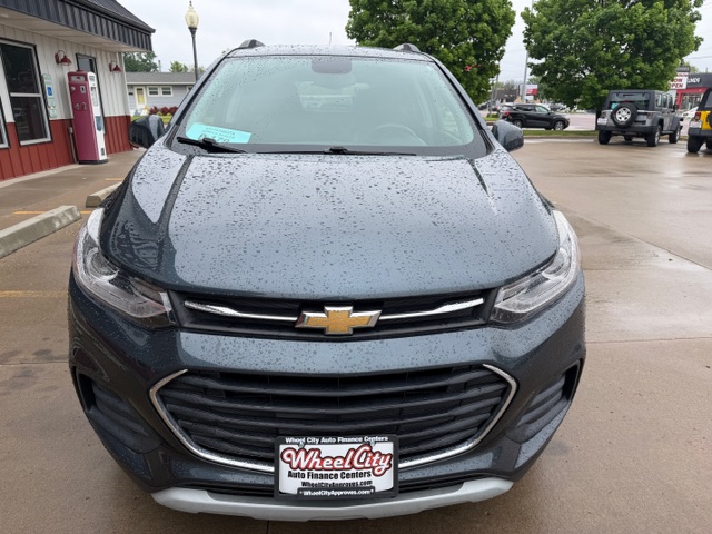 Front view of a dark gray Chevrolet SUV with raindrops on the hood, parked in a dealership lot near a red building; license plate frame reads Wheel City Auto Finance Centers.