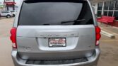 Rear view of a silver Dodge minivan with a Wheel City dealer plate, parked in a dealership lot under a cloudy sky.