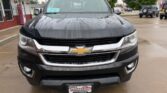 Front view of a black Chevrolet pickup truck parked in a dealership lot on a rainy day, showing the chrome grille, gold bowtie emblem, and Wheel City plate.