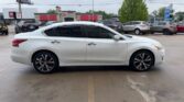 White four-door sedan in a dealership lot, side view with dark tinted windows and black alloy wheels, parked among other cars