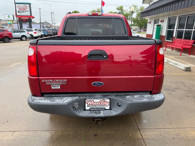 Rear view of a red Ford Explorer pickup with the tailgate up, chrome bumper, and a dealership license plate frame on a wet parking lot surface.