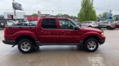 Red pickup truck with a double cab parked in a car dealership lot with other vehicles around and a tree in the background.