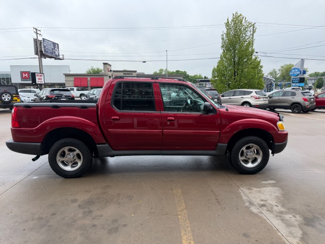Red pickup truck with a double cab parked in a car dealership lot with other vehicles around and a tree in the background.