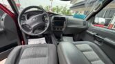 Inside a grey pickup truck, with the driver's seat and front passenger seat visible, steering wheel on the left and a center dashboard/radio, viewed from the rear seats toward a dealership exterior.
