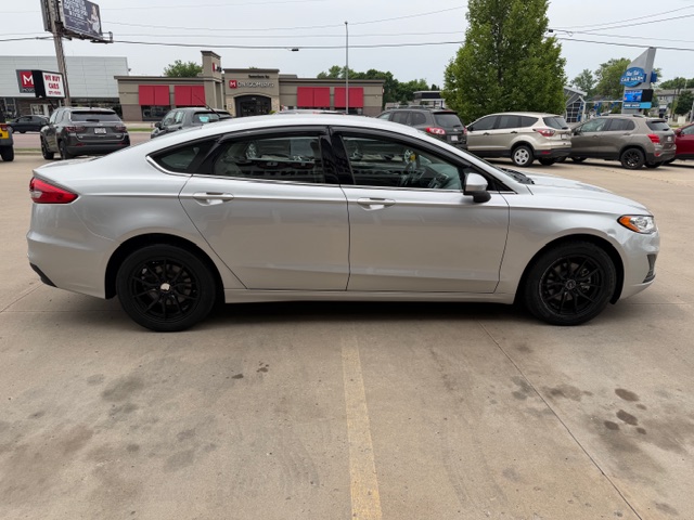Side view of a silver four-door sedan with black wheels parked in a car lot outside a dealership.