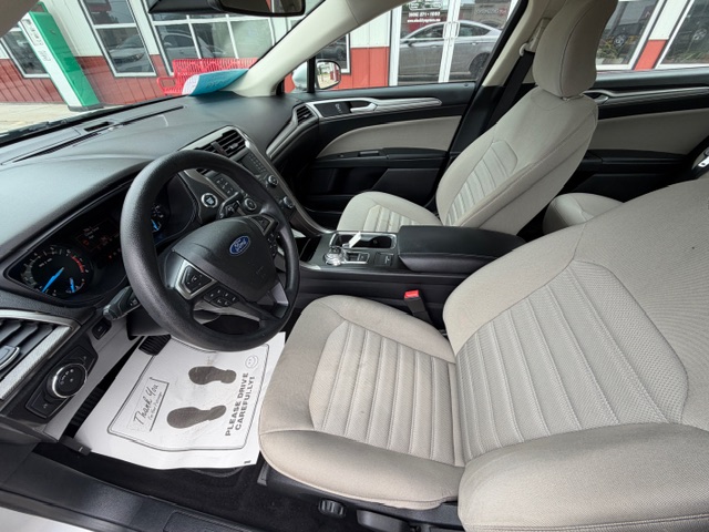 Front interior of a Ford SUV with beige fabric seats, black dashboard, and steering wheel; driver’s seat shows controls and center console; paper floor mat with footprints on the driver side.