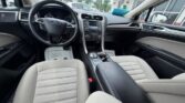 Interior of a Ford sedan from the front passenger side, showing beige cloth seats, center console, steering wheel, and a 'Thank you' mat on the driver's floor.