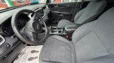 Interior view of a gray-car front cabin showing steering wheel, dashboard, center console, and gray cloth seats with a factory floor mat visible on the driver’s side.