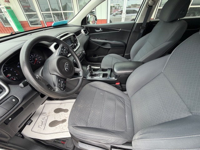 Interior view of a gray-car front cabin showing steering wheel, dashboard, center console, and gray cloth seats with a factory floor mat visible on the driver’s side.