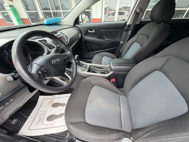 Front interior of a Kia car showing the steering wheel, dashboard with center console, gear shifter, and two-tone fabric seats; floor mat visible.