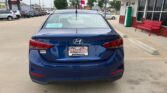 Blue Hyundai Accent parked in a dealership lot, rear view, with Wheel City Auto Finance plate frame and a red bench nearby on the right.