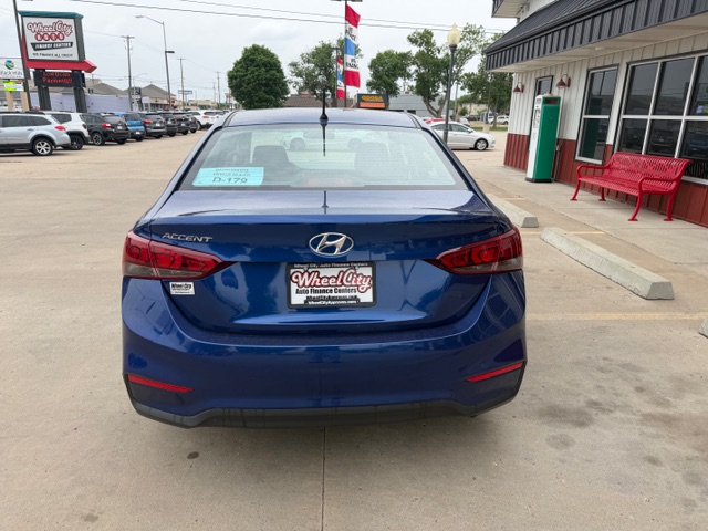 Blue Hyundai Accent parked in a dealership lot, rear view, with Wheel City Auto Finance plate frame and a red bench nearby on the right.