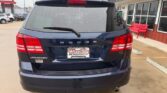 Rear view of a blue Dodge Journey SUV in a dealership lot, with a Wheel City license plate frame and a wet rear window.