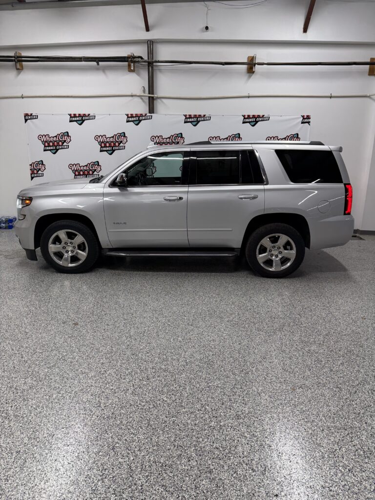 Side view of a silver Chevrolet Tahoe SUV parked indoors, with a Wheel City Auto Finance Centers banner in the background.