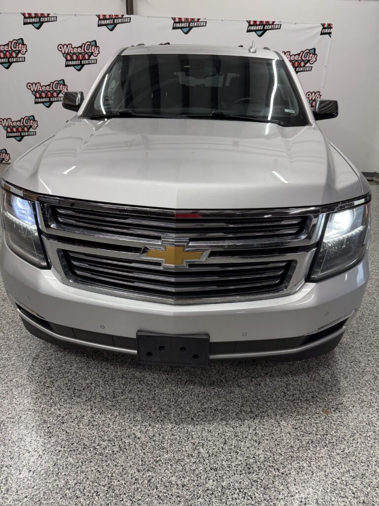 Front view of a silver Chevrolet SUV in a dealership showroom, with headlights on and a chrome grille featuring the gold bowtie emblem.