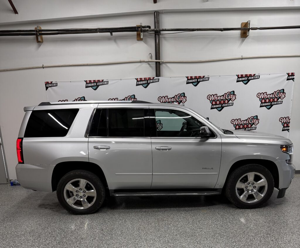Side profile of a silver SUV (Chevrolet Tahoe) parked indoors, with a Wheel City Auto Finance Centers banner on the wall behind it.