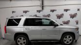 Side profile of a silver SUV (Chevrolet Tahoe) parked indoors, with a Wheel City Auto Finance Centers banner on the wall behind it.