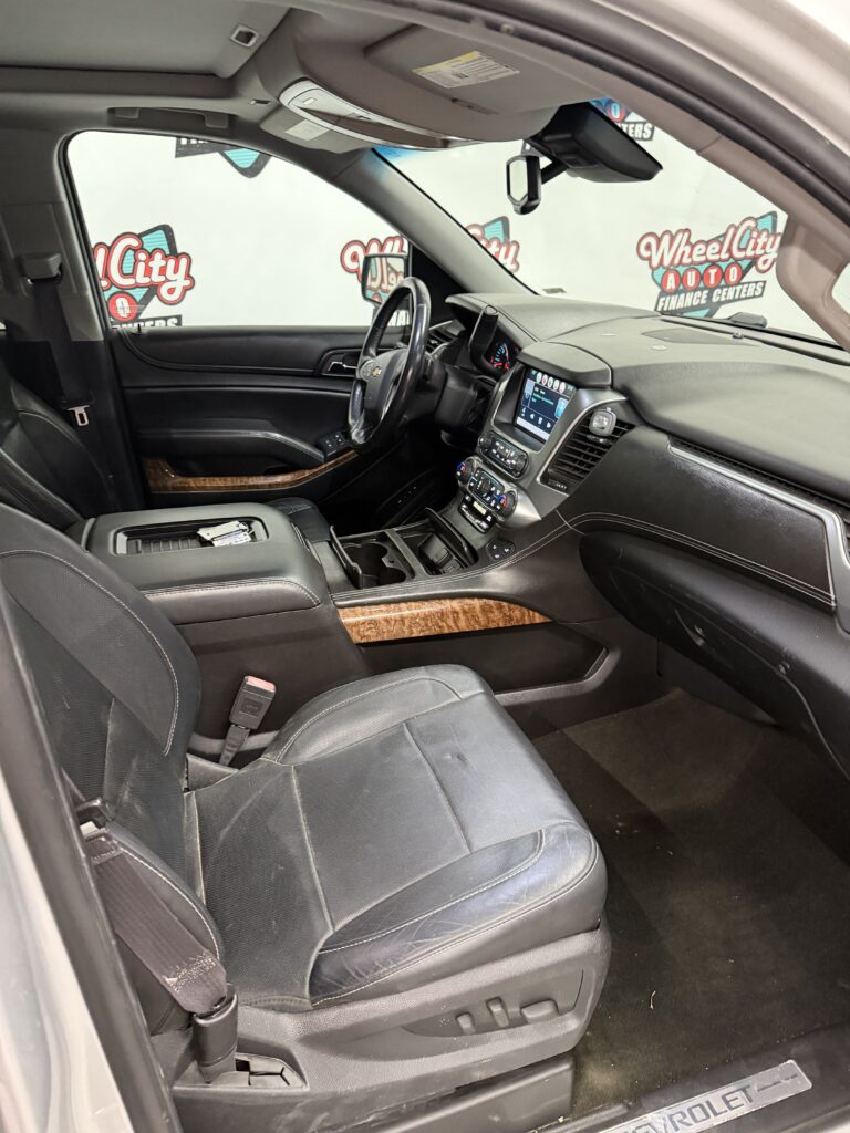 Front interior of a black Chevrolet SUV showing the driver’s seat, steering wheel, center console, and wood trim accents in a showroom setting.