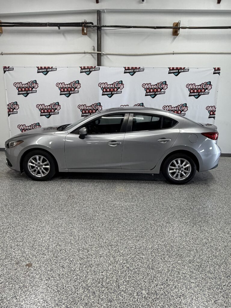Side profile of a silver sedan parked indoors with a backdrop displaying Wheel City Auto Finance Centers logos