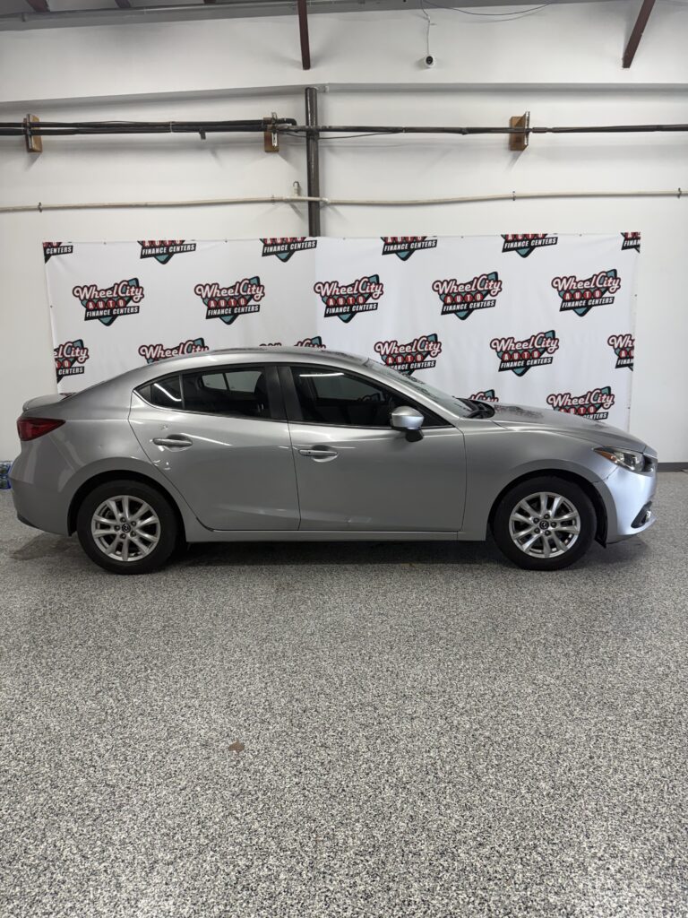 Side view of a silver four-door sedan parked indoors in front of a white backdrop with Wheel City Auto Finance Centers logos.