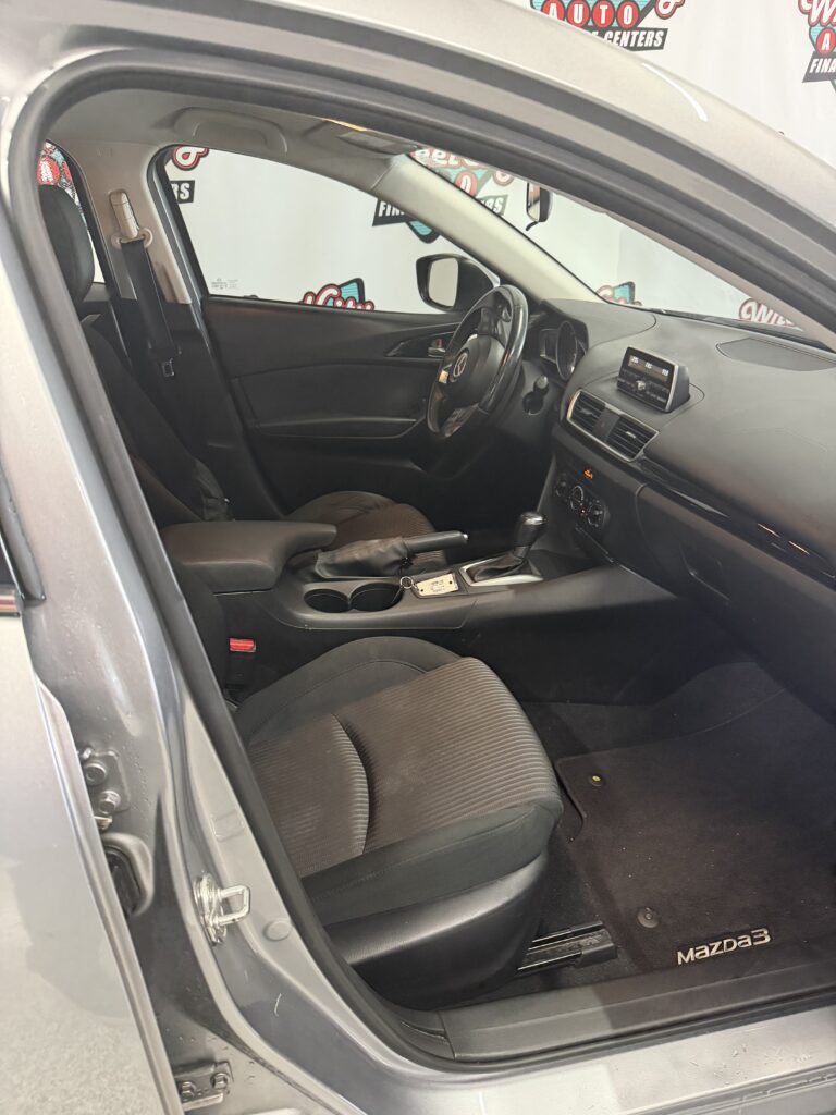 Interior of a Mazda3 showing the front seats, steering wheel, center console, and dashboard viewed from the open driver's side door.