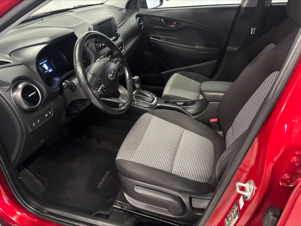 Interior of a modern car cabin: driver's seat, steering wheel, center touchscreen, and front passenger seat visible in a red vehicle door frame