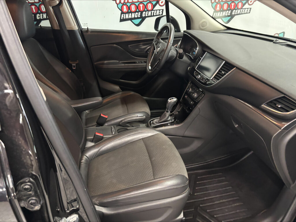 Interior view of a black leather car cabin with the steering wheel, center console, and touchscreen display visible; dealership signage seen through the windows in the background.