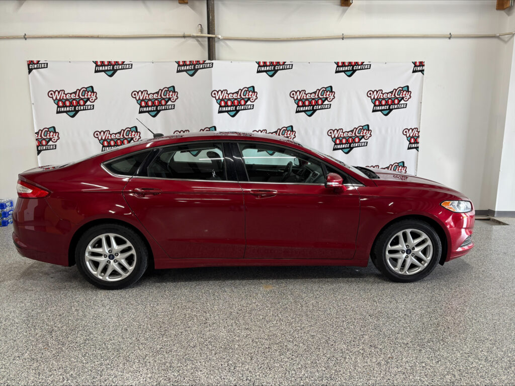 Red sedan parked indoors in front of a white wall banner that reads Wheel City Auto Finance Centers repeatedly.